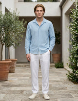 Man wearing a light blue shirt and white pants standing in an outdoor setting with plants and a building.