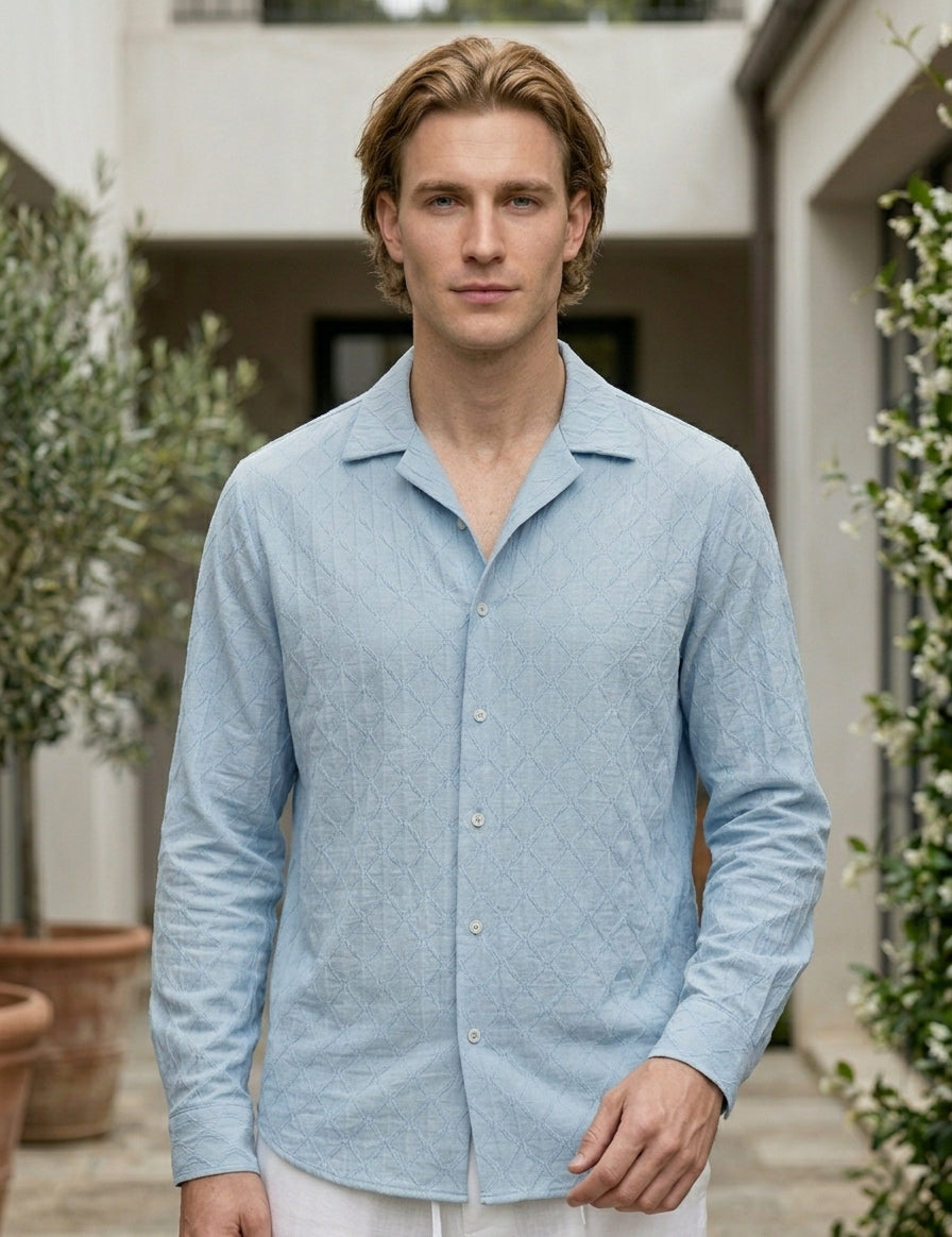 Man wearing a light blue shirt standing outdoors with greenery and a building in the background