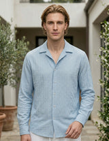 Man wearing a light blue shirt standing outdoors with greenery and a building in the background