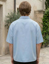 Man wearing a light blue shirt standing outdoors with a building and greenery in the background