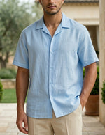 Man wearing a light blue shirt standing outdoors with greenery and a stone building in the background