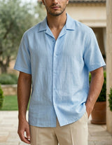 Man wearing a light blue shirt standing outdoors with greenery and a stone building in the background