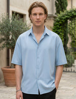 Man wearing a light blue shirt standing outdoors with plants in the background