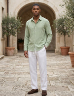 Man wearing a light green shirt and white pants standing in an outdoor setting with potted plants.