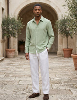 Man wearing a light green shirt and white pants standing in an outdoor setting with potted plants.