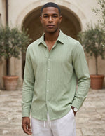 Man wearing a light green shirt and white pants standing outdoors with a stone archway and plants in the background.