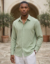 Man wearing a light green shirt and white pants standing outdoors with a stone archway and plants in the background.