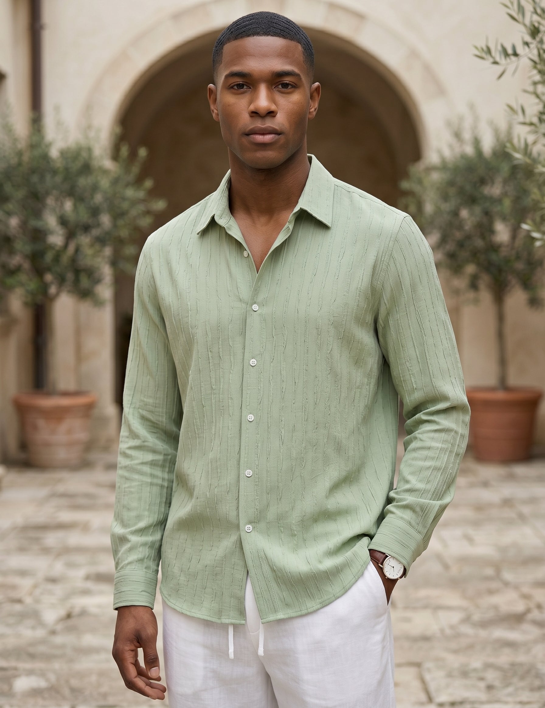 Man wearing a light green shirt and white pants standing outdoors with a stone archway and plants in the background.