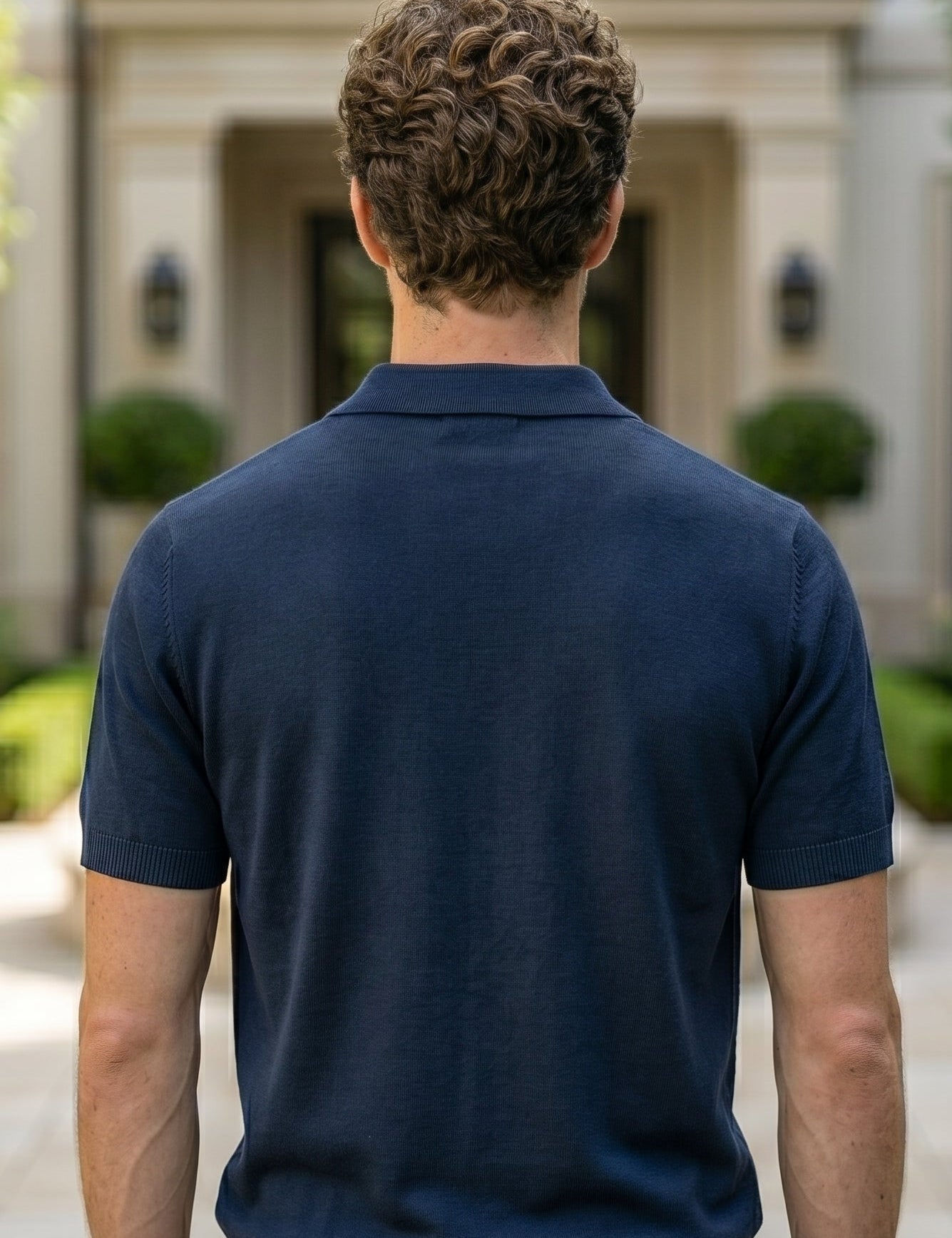 Man wearing a navy blue polo shirt standing in front of a building with greenery.