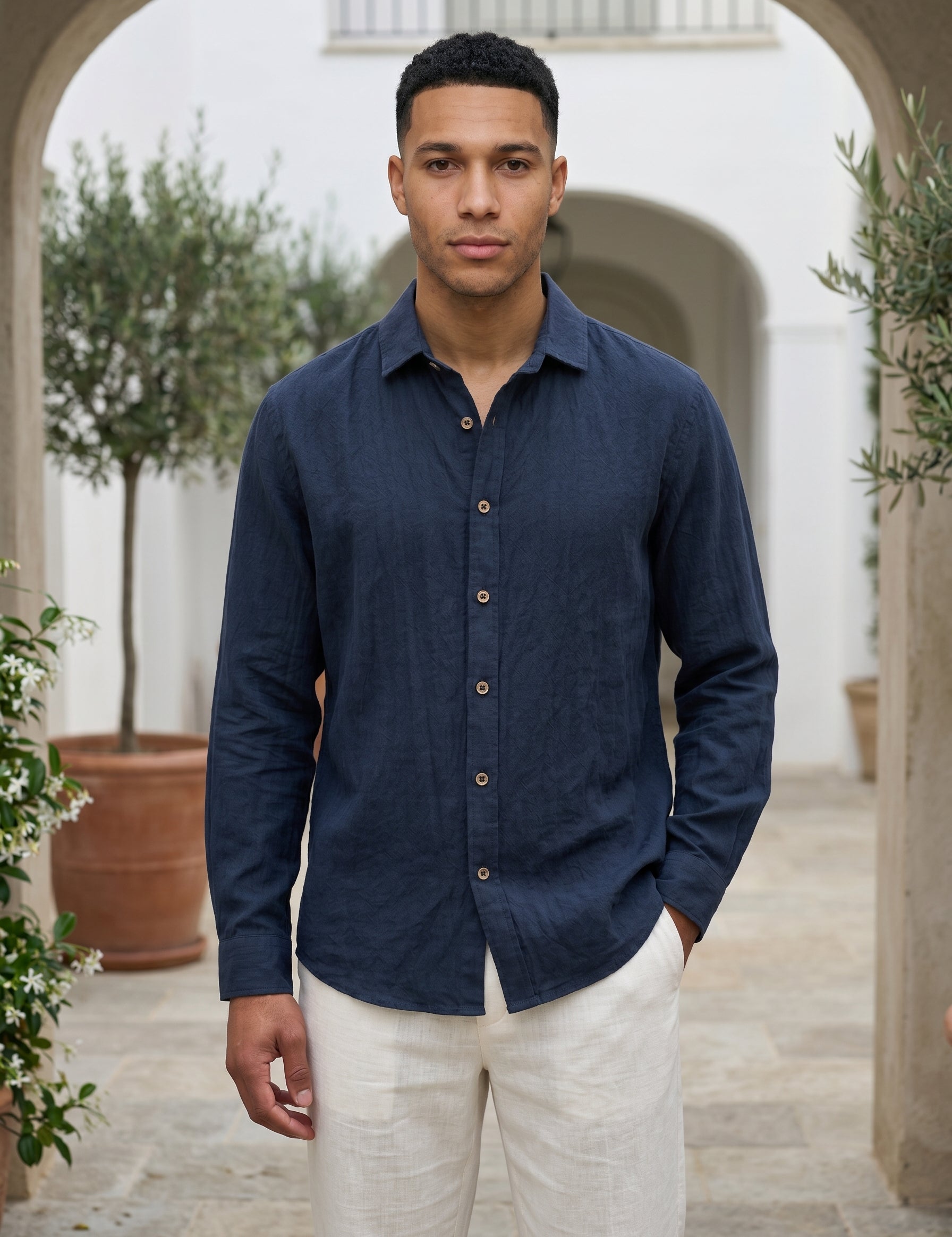Man wearing a navy blue shirt and white pants standing in an outdoor setting with arches and plants.