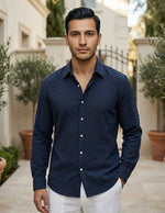 Man wearing a navy blue shirt and white pants standing outdoors with plants and a building in the background