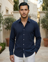 Man wearing a navy blue shirt and white pants standing outdoors with plants and a building in the background