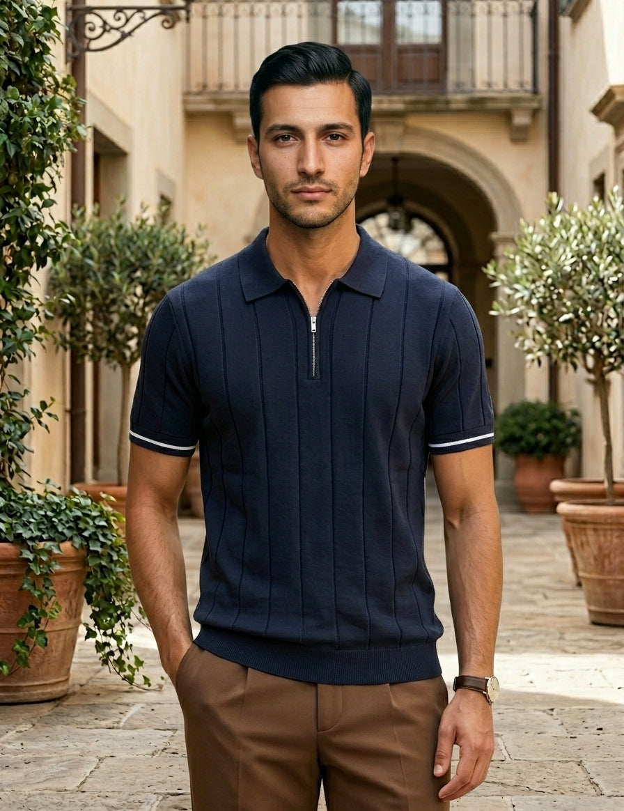 Man wearing a navy polo shirt and brown pants standing in an outdoor setting with plants and a building.