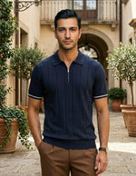 Man wearing a navy polo shirt and brown pants standing in an outdoor setting with plants and a building.