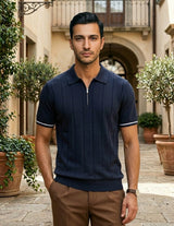 Man wearing a navy polo shirt and brown pants standing in an outdoor setting with plants and a building.