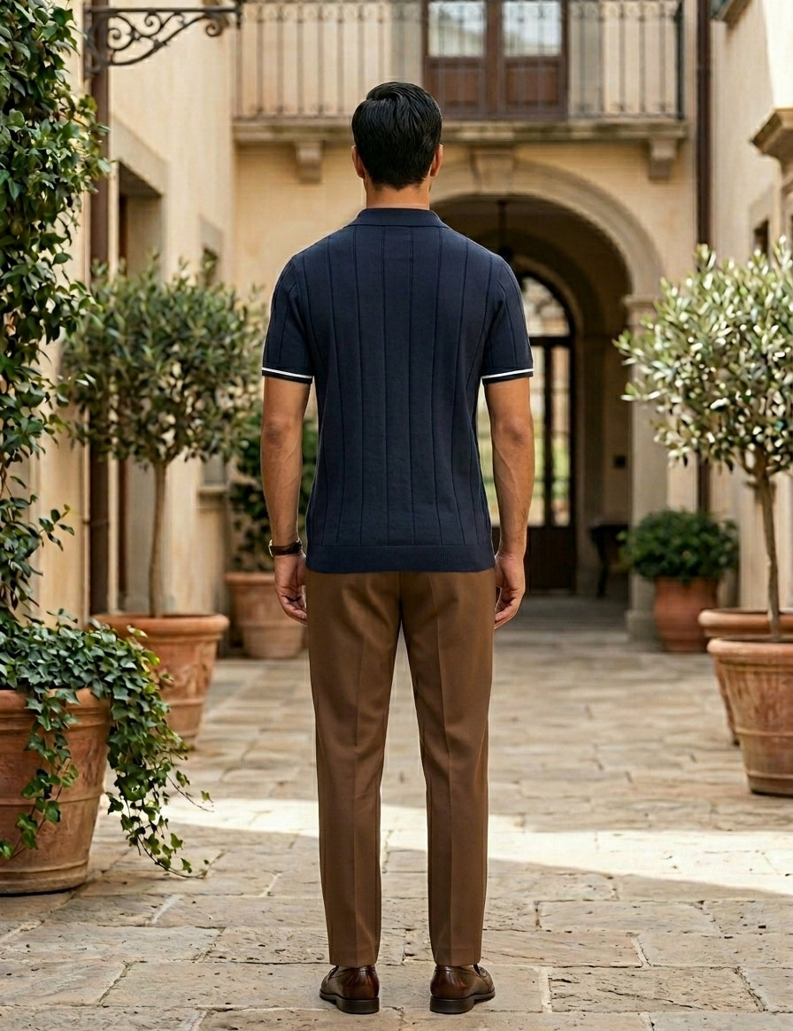 Man wearing a navy polo shirt and brown pants standing in an outdoor setting with potted plants.