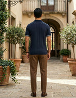 Man wearing a navy polo shirt and brown pants standing in an outdoor setting with potted plants.