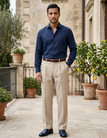 Man wearing a navy shirt and beige pants standing in an outdoor setting with plants and stone walls.