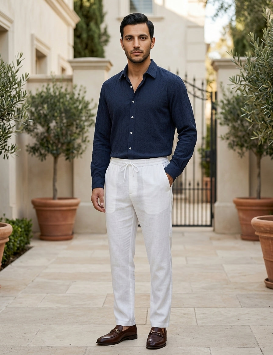 Man wearing a navy shirt and white pants standing in an outdoor setting with potted plants.