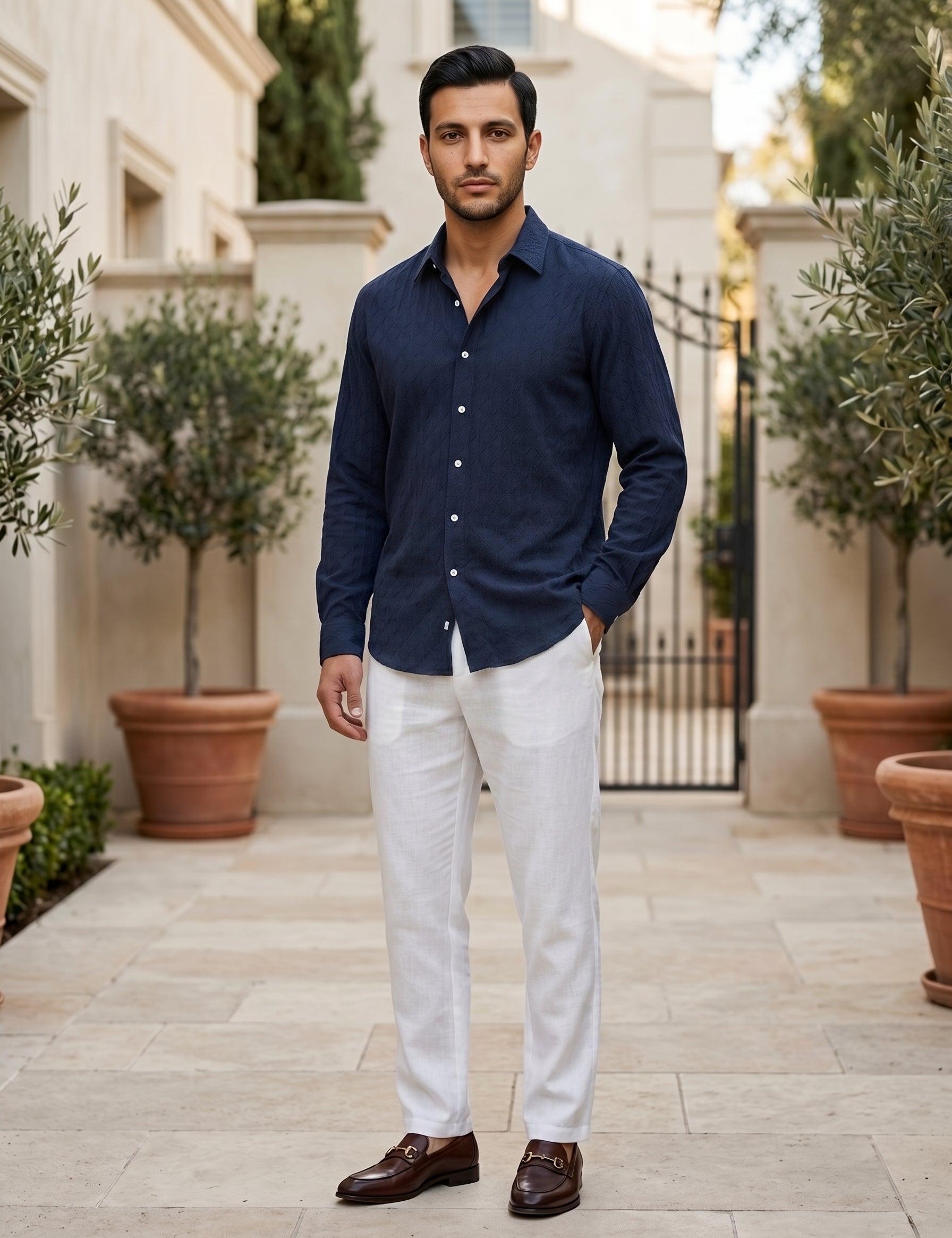 A man wearing a navy blue long-sleeve button-down shirt, white trousers, and brown leather loafers, standing in a paved outdoor courtyard with potted olive trees.