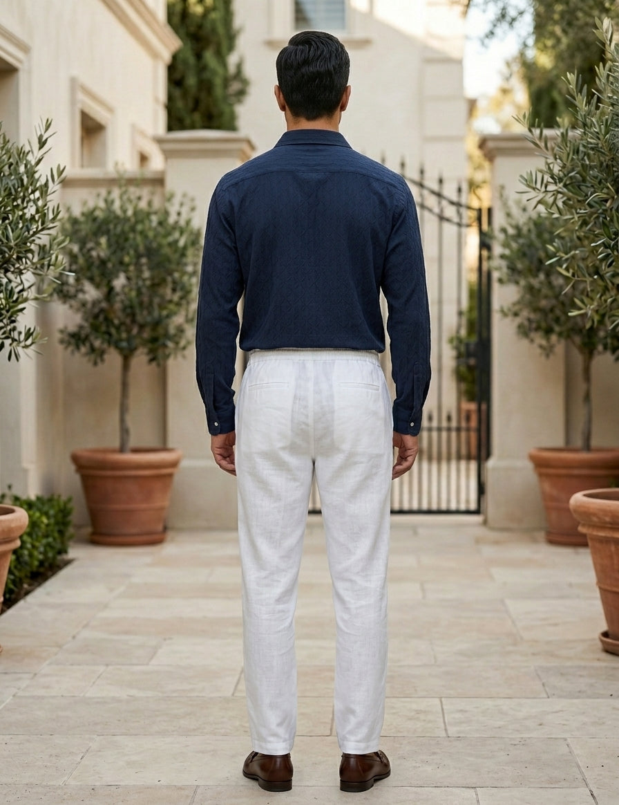 Man wearing a navy shirt and white pants standing in an outdoor setting with potted plants.