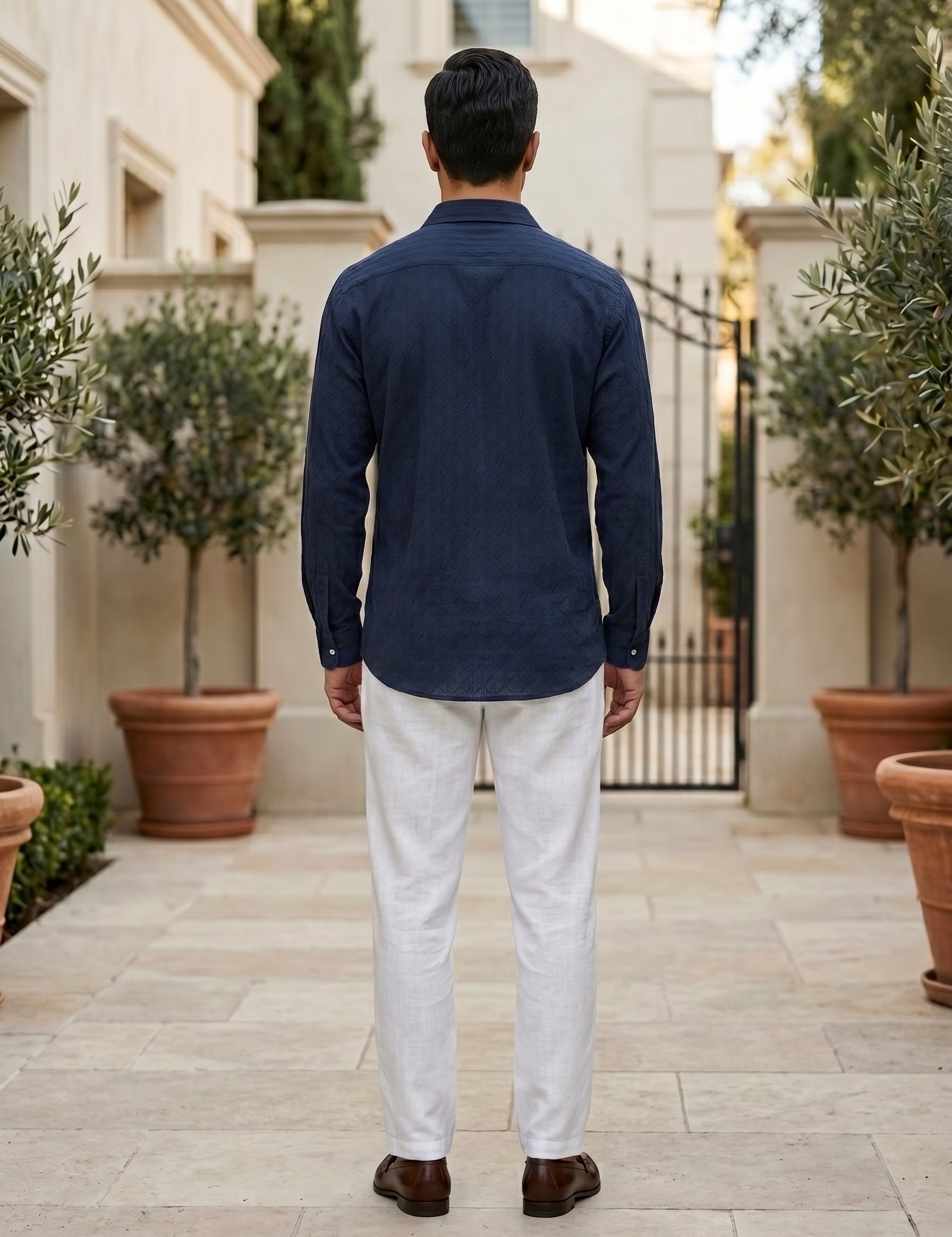 Man wearing a navy shirt and white pants standing in an outdoor setting with potted plants.