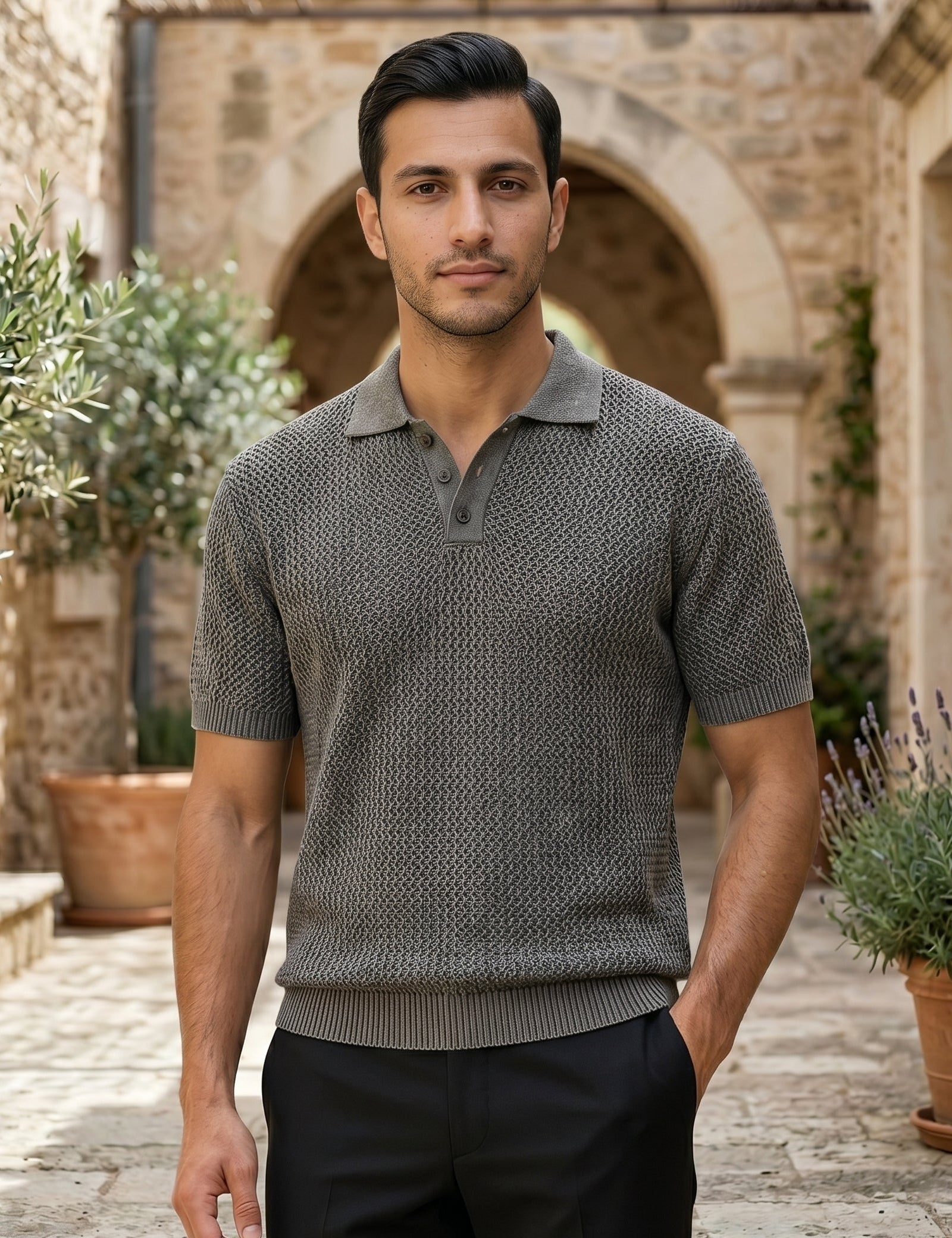 Man wearing a patterned short-sleeve shirt standing in an outdoor setting with stone architecture and plants.