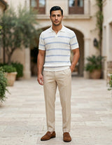Man wearing a striped polo shirt and beige pants standing in an outdoor setting with plants and buildings.