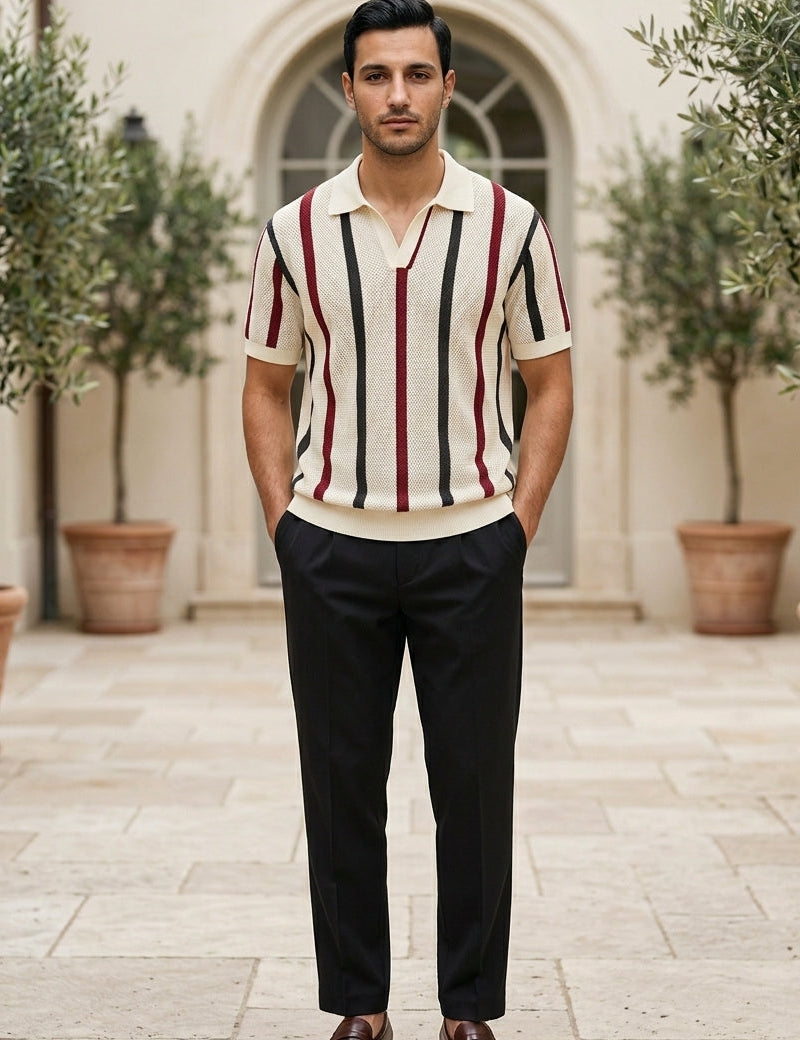 Man wearing a striped polo shirt and dark pants standing outdoors with plants and a building in the background.