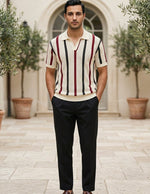 Man wearing a striped polo shirt and dark pants standing outdoors with plants and a building in the background.