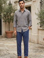 Man wearing a striped shirt and blue pants standing in an outdoor setting with stone pavement and plants.