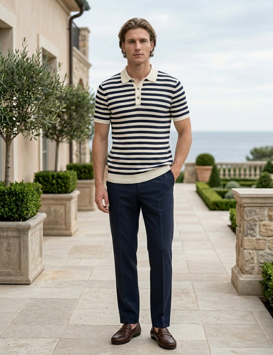 Man wearing a striped shirt and dark pants standing in an outdoor setting with a building and garden in the background.