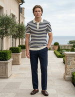 Man wearing a striped shirt and dark pants standing in an outdoor setting with a building and garden in the background.
