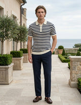 Man wearing a striped shirt and dark pants standing in an outdoor setting with a building and garden in the background.