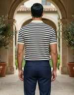 Man wearing a striped shirt and dark pants standing in an outdoor setting with arches and plants.