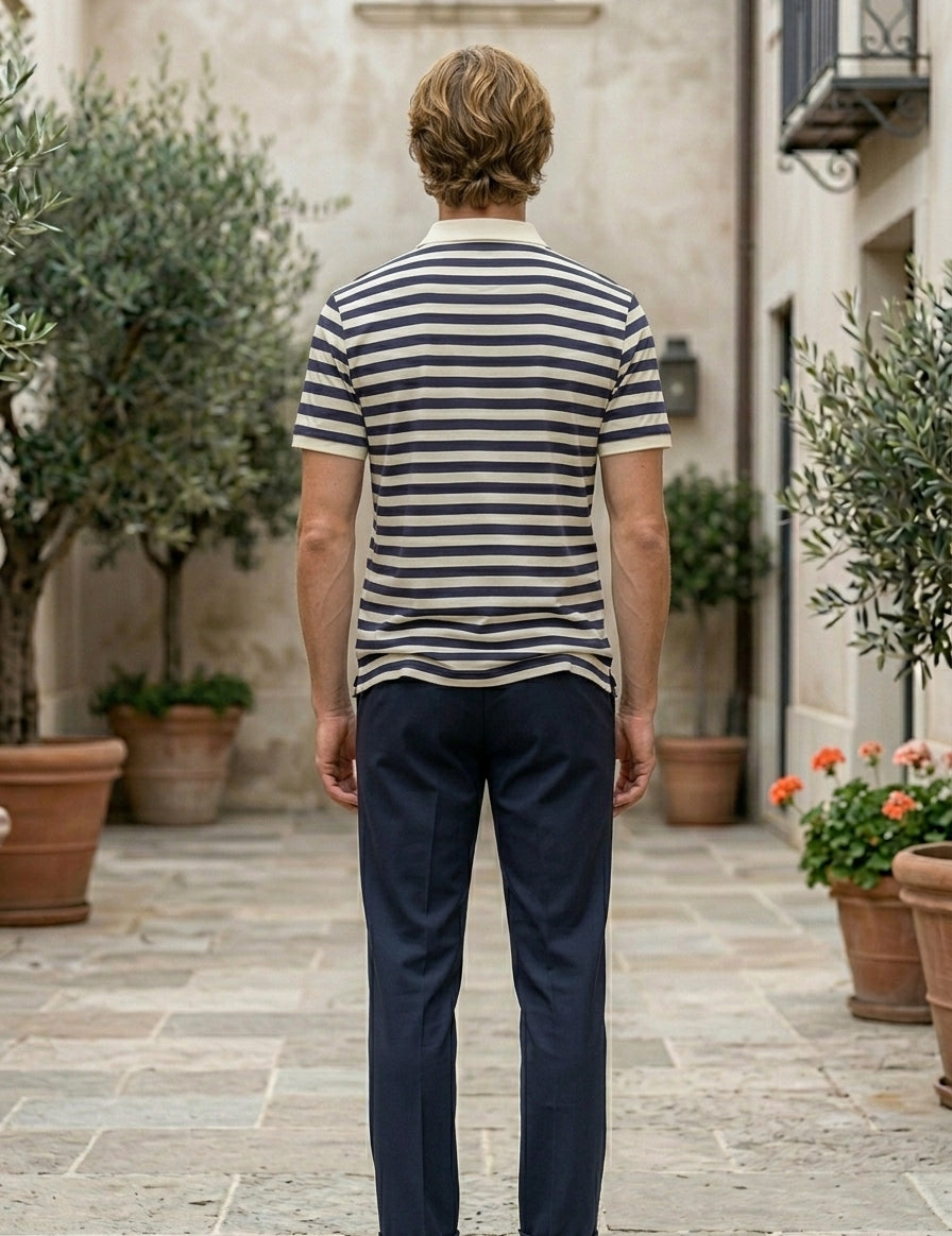 Man wearing a striped shirt and dark pants standing in an outdoor setting with plants and buildings.