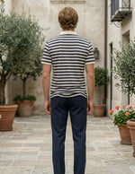 Man wearing a striped shirt and dark pants standing in an outdoor setting with plants and buildings.