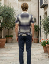 Man wearing a striped shirt and dark pants standing in an outdoor setting with plants and buildings.