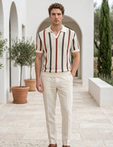 Man wearing a striped shirt and white pants standing in a sunlit outdoor setting with plants and arches.