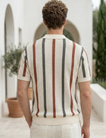 Man wearing a striped shirt and white pants standing in an outdoor setting with arches and plants.