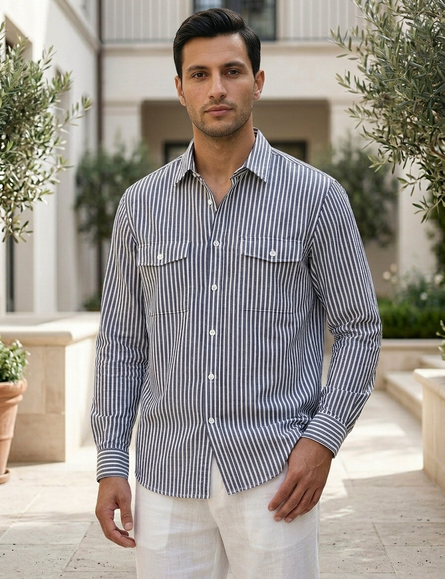 Man wearing a striped shirt and white pants standing outdoors with greenery and a building in the background