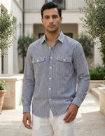 Man wearing a striped shirt and white pants standing outdoors with greenery and a building in the background