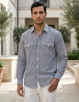 Man wearing a striped shirt and white pants standing outdoors with greenery and a building in the background