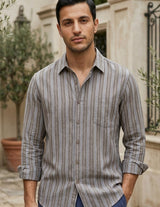 Man wearing a striped shirt standing outdoors with plants and a building in the background