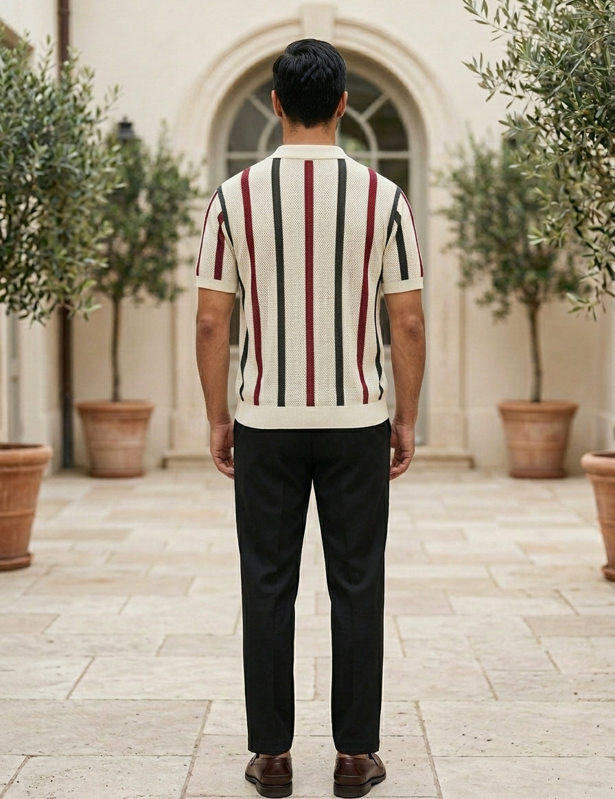 Man wearing a striped sweater and dark pants standing in an outdoor setting with potted plants.