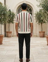 Man wearing a striped sweater and dark pants standing in an outdoor setting with potted plants.