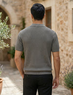 Man wearing a textured gray sweater walking outdoors with stone architecture and plants in the background