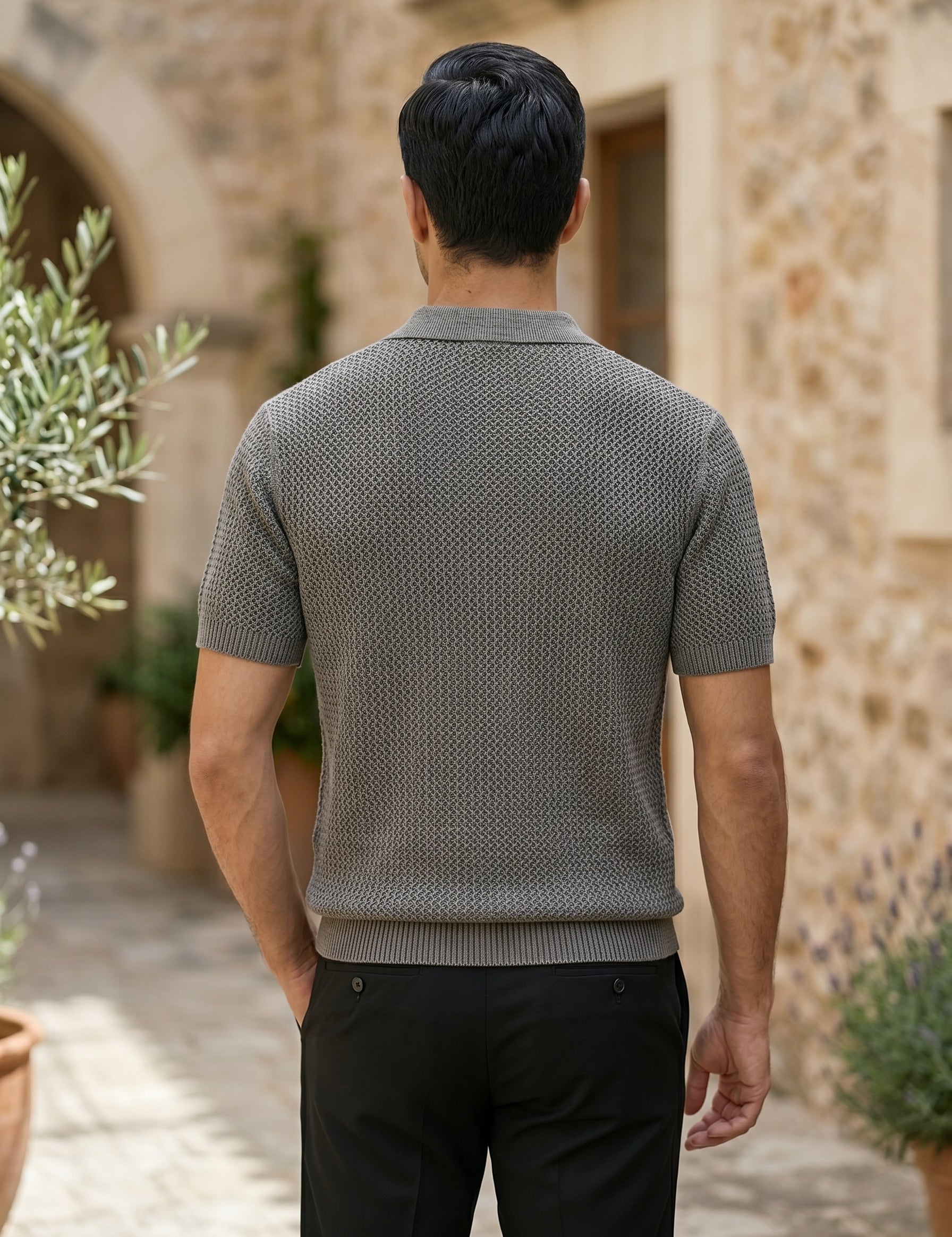 Man wearing a textured gray sweater walking outdoors with stone architecture and plants in the background