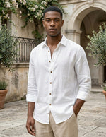 Man wearing a white button-up shirt and beige pants standing in an outdoor setting with architectural elements and plants.