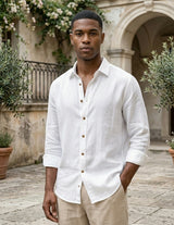 Man wearing a white button-up shirt and beige pants standing in an outdoor setting with architectural elements and plants.